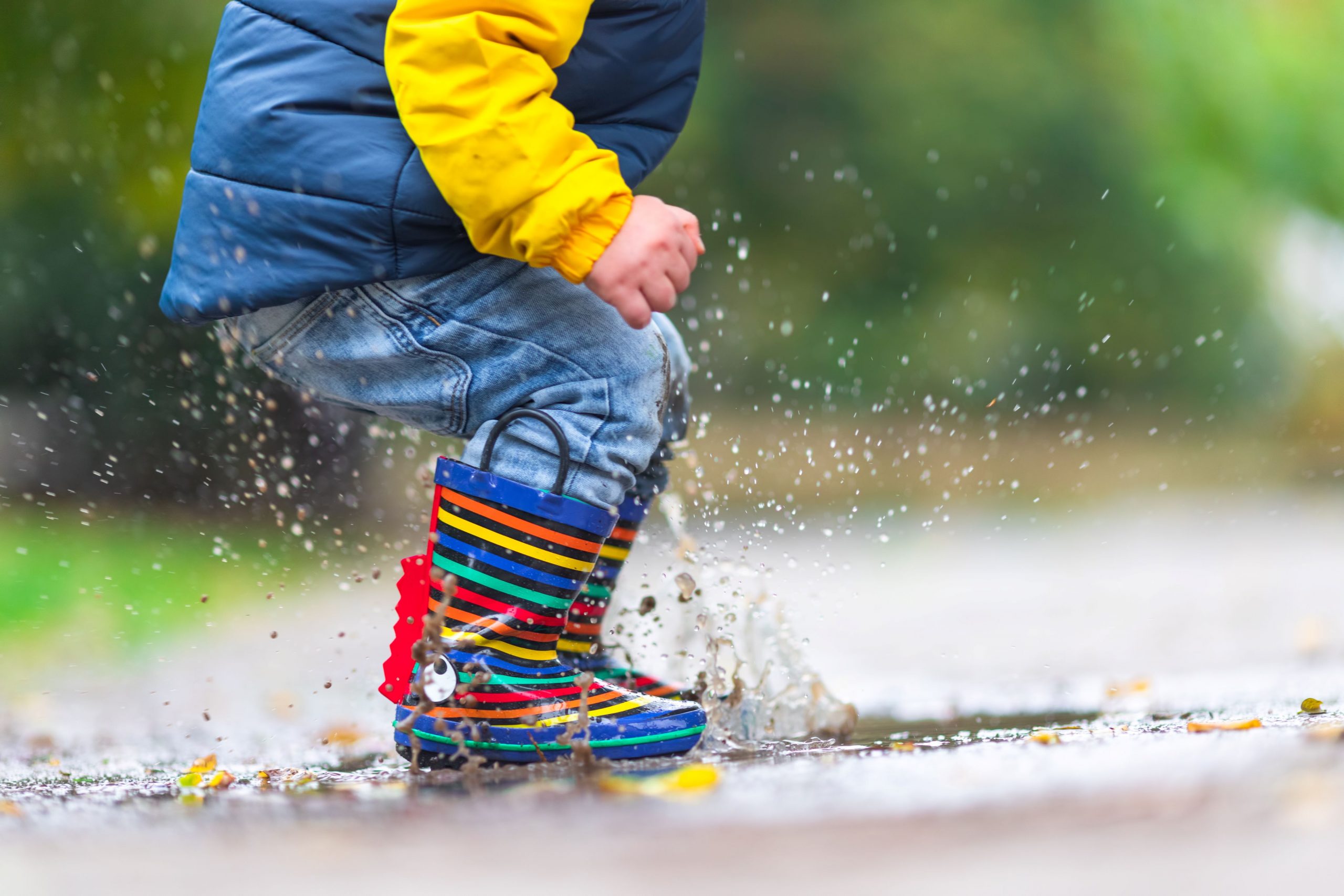 A child wearing rainbow wellies and a blue and yellow raincoat jimps in a puddle, splashing water around